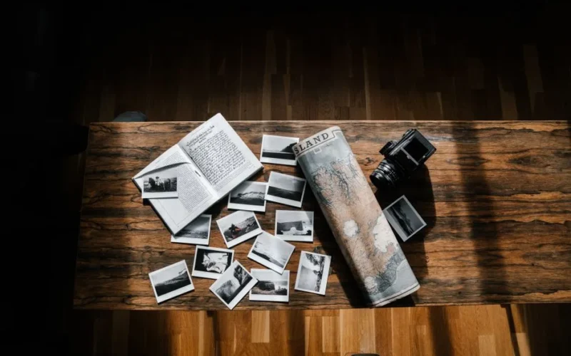 Composition of black and white photos and old travel log placed with vintage island map and obsolete photo camera on narrow wooden table