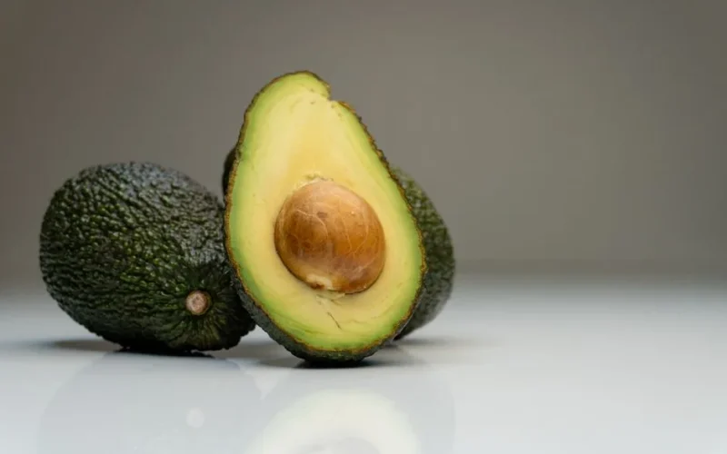 Close-up of ripe avocados with a sliced half showing the seed, against a gray background.