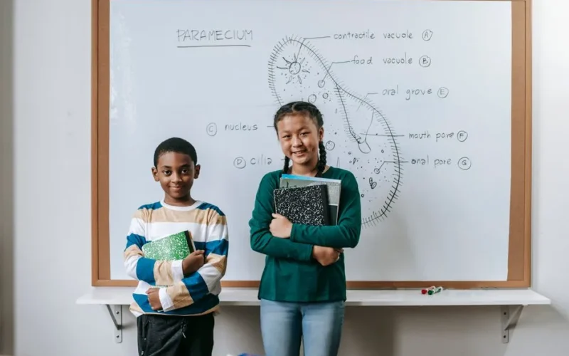 Cheerful little multiethnic students standing near whiteboard with books and looking at camera during biology lesson