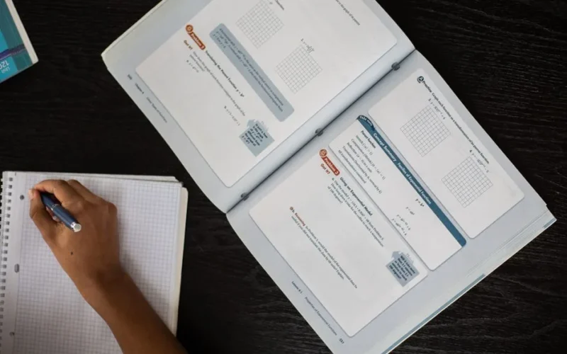 Top view of faceless African American student taking notes in notepad while sitting at table with textbook during lesson in class