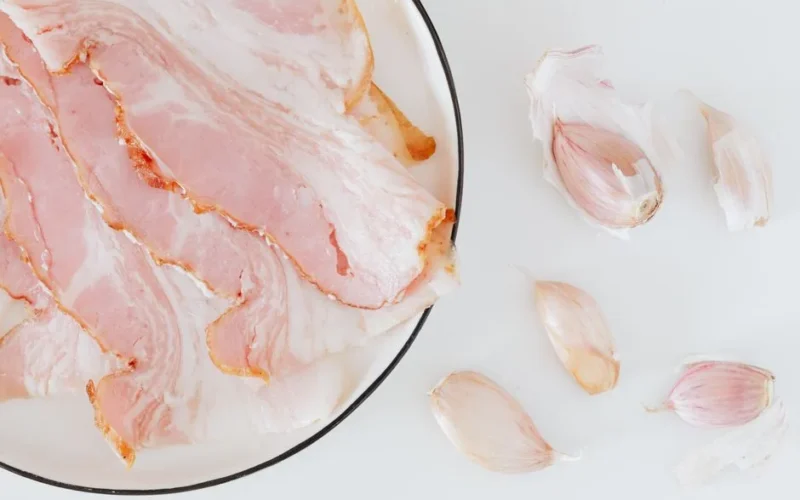 Top view of plate with bacon slices and pieces of garlic placed on white surface