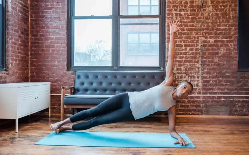 Full body of active African American female doing Vasisthasana posture on mat during yoga training at home with brick walls