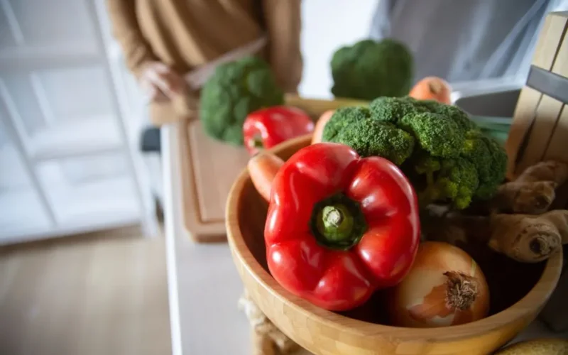 A close-up of fresh vegetables including red peppers, broccoli, and ginger in a wooden bowl.