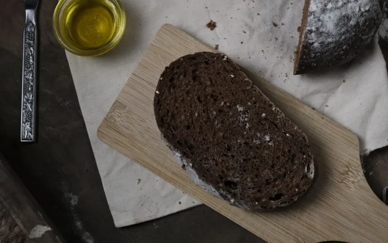 A slice of rustic whole wheat bread on a wooden cutting board with olive oil on the side.
