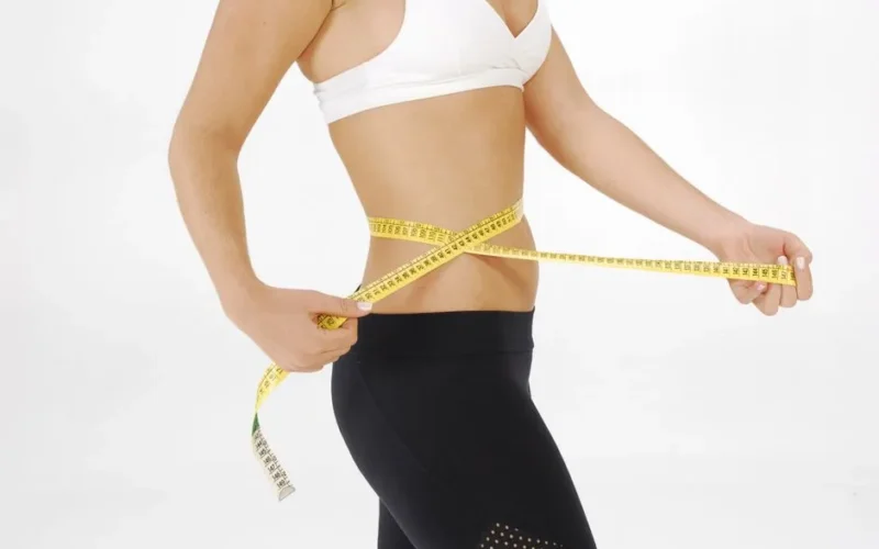 Fit woman in sportswear measuring her waist with a tape measure against a white background.