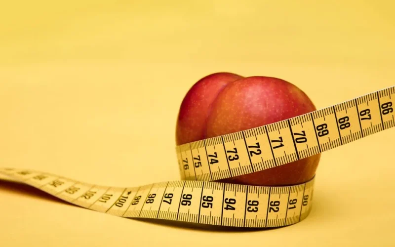 A peach wrapped with a measuring tape on a vibrant yellow background, symbolizing health and measurement.
