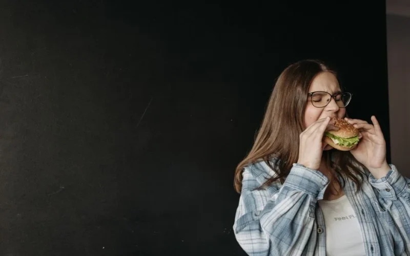 Woman in casual attire eating a hamburger in front of a dark background.
