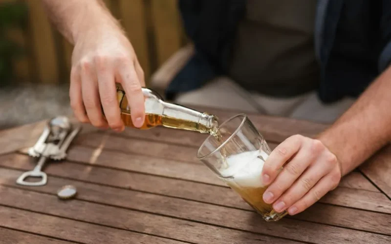 From above of crop anonymous male sitting at wooden table and filling glass with beer