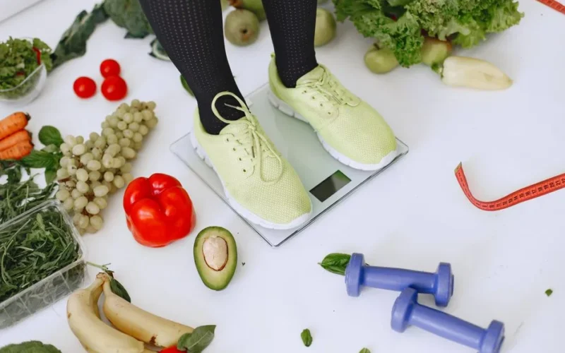 Person standing on scales surrounded by fresh produce and fitness equipment symbolizing a balanced lifestyle.