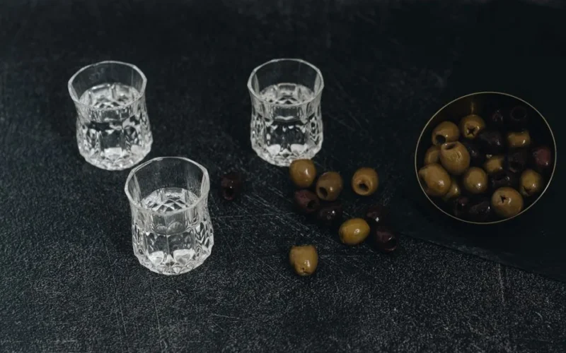 Top view of crystal glasses filled with vodka and bowl of olives on a dark textured surface.