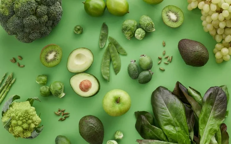 Top view of assorted fresh green vegetables and fruits on a green background, perfect for healthy lifestyle visuals.