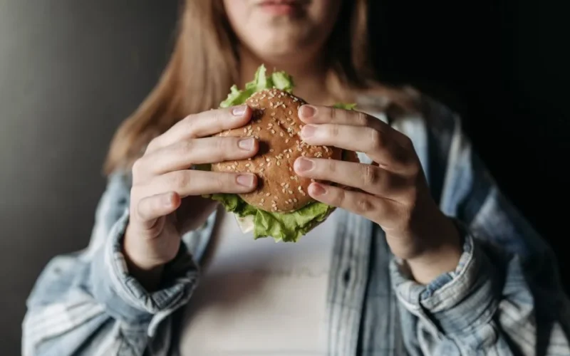A person in a casual shirt holding a fresh burger with lettuce and sesame bun.