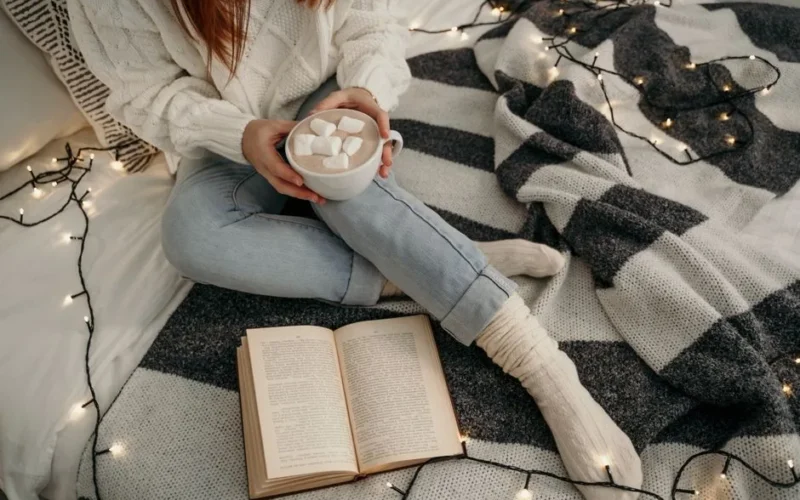 A woman enjoys marshmallow hot chocolate, sitting on a bed with fairy lights and a book, exuding cozy winter vibes.