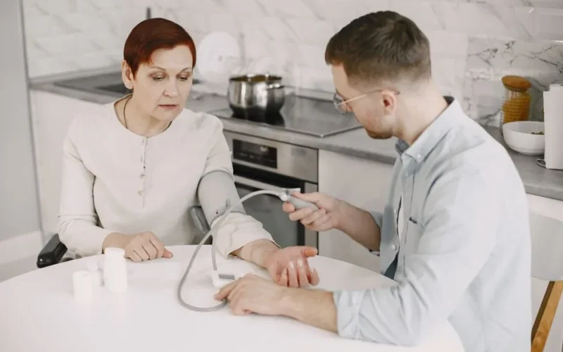 Adult man monitoring elderly woman's blood pressure at kitchen table using sphygmomanometer.