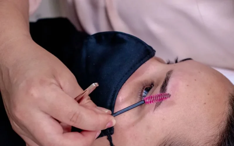 Close-up of a beautician performing eyebrow grooming on a client wearing a mask.