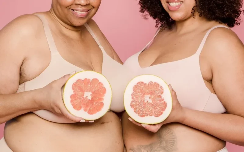 Anonymous plus size African American female models in lingerie with halves of ripe grapefruit in hands smiling on pink background in studio