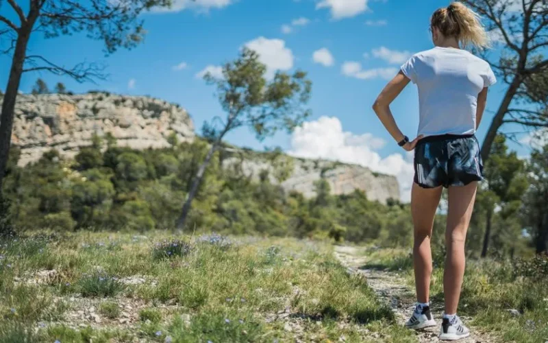 A woman explores a mountain trail surrounded by nature during a sunny day.