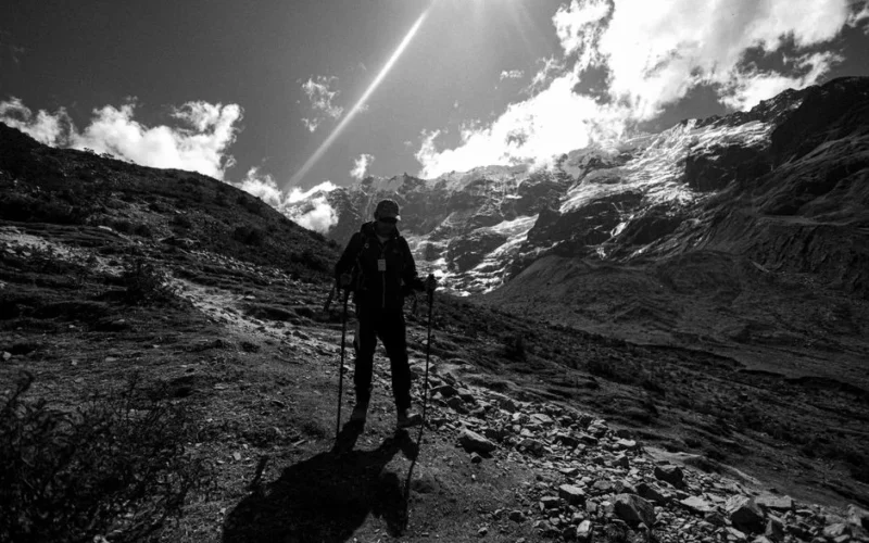 Silhouette of a hiker with poles on a rocky mountain path under a bright, dramatic sky.
