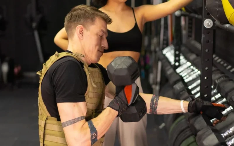 Side view of a man working out with a dumbbell and weighted vest in a gym setting.