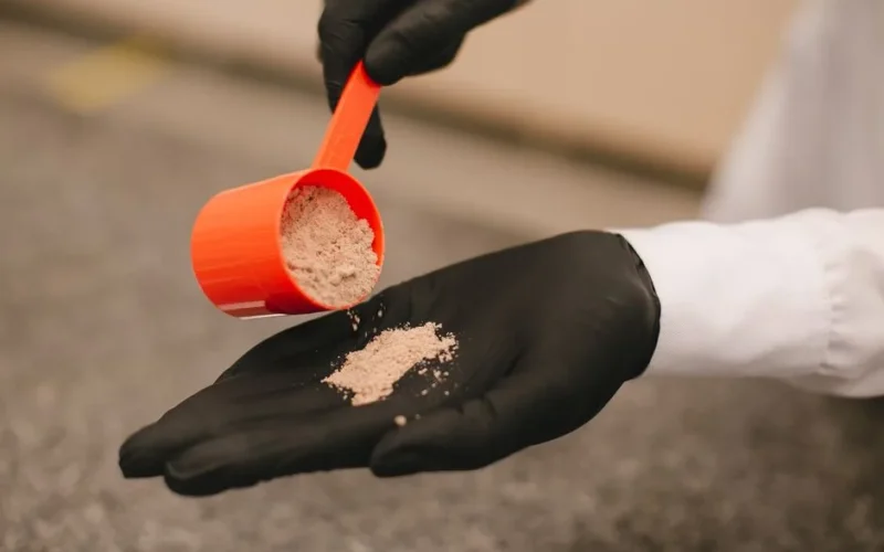 A close-up shot of a black-gloved hand pouring powder from a red scoop, over a textured surface.