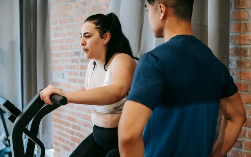 Side view of serious concentrated young obese female doing weight lose exercise on cycling equipment during cardio training with crop ethnic male personal coach