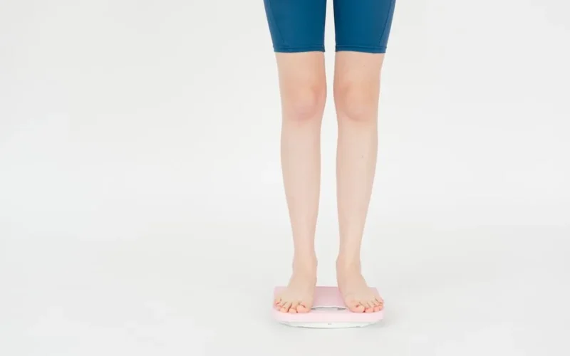 Crop anonymous female in sportswear measuring weight on scales for body control against white background