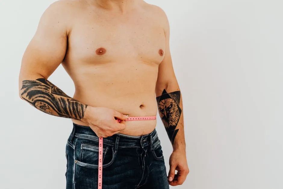 Shirtless man using measuring tape to check waist size, showcasing tattoos for fitness concept.