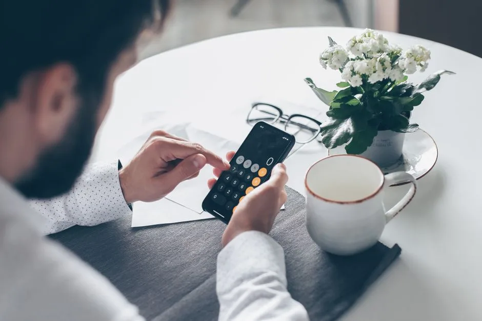 Adult man using smartphone calculator at table with flowers and coffee cup.