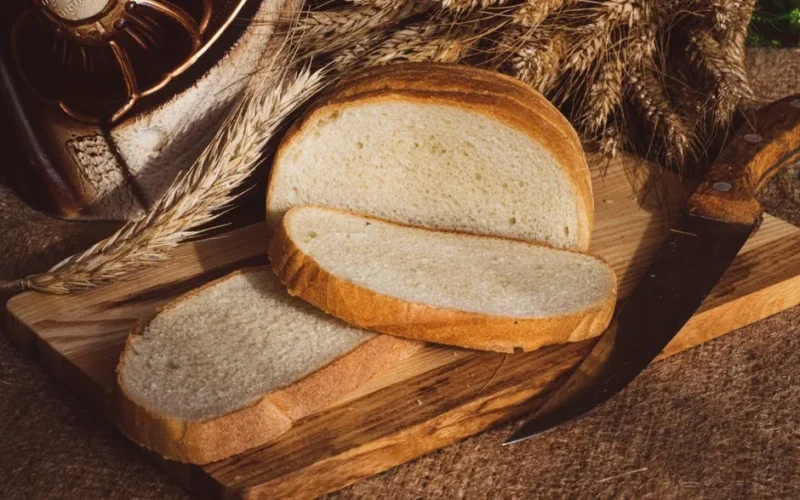 Freshly sliced bread on a rustic wooden board with wheat and a knife.
