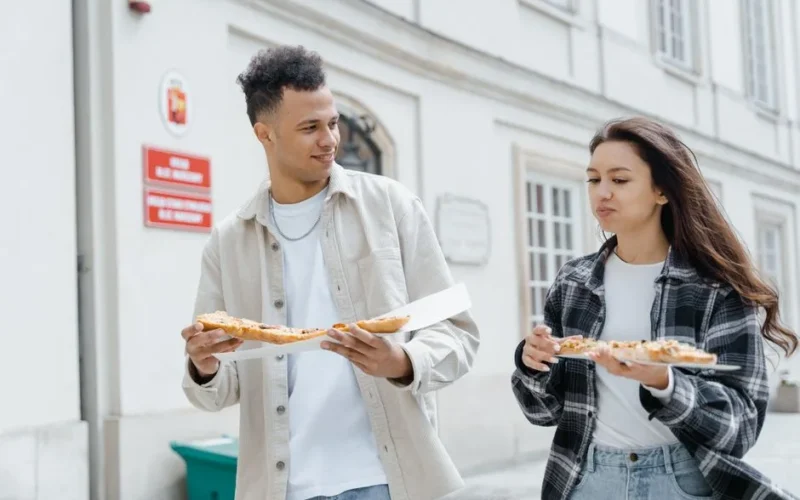 Two young adults enjoying pizza while walking outdoors in the city, creating a joyful street food moment.