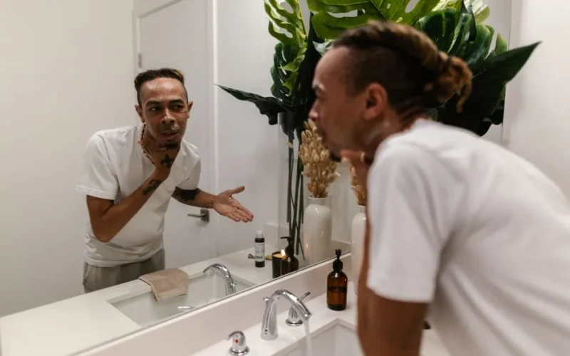 A young man applying skincare product while looking in the mirror in a modern bathroom.