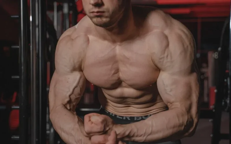 Shirtless man displaying muscular physique with focus on biceps and abs in a gym.