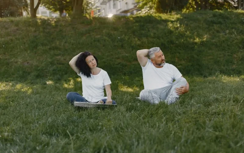 Couple doing neck stretches outdoors, promoting a healthy lifestyle in a sunny park setting.