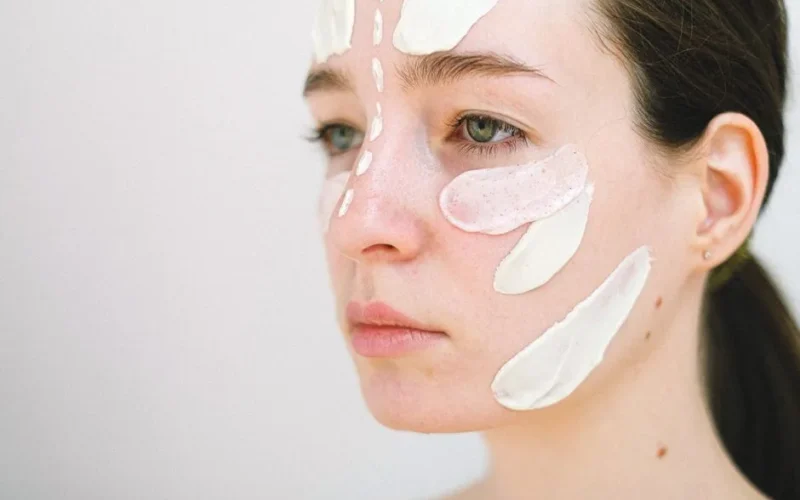 Side view of crop female model spreading white cream on face during skincare procedure on white background