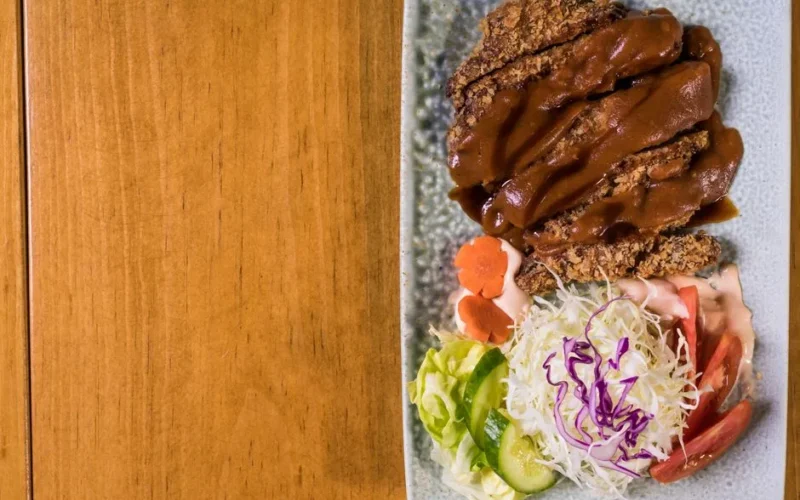 Delicious breaded pork cutlet drizzled with sauce, accompanied by a fresh vegetable salad on a wooden surface.