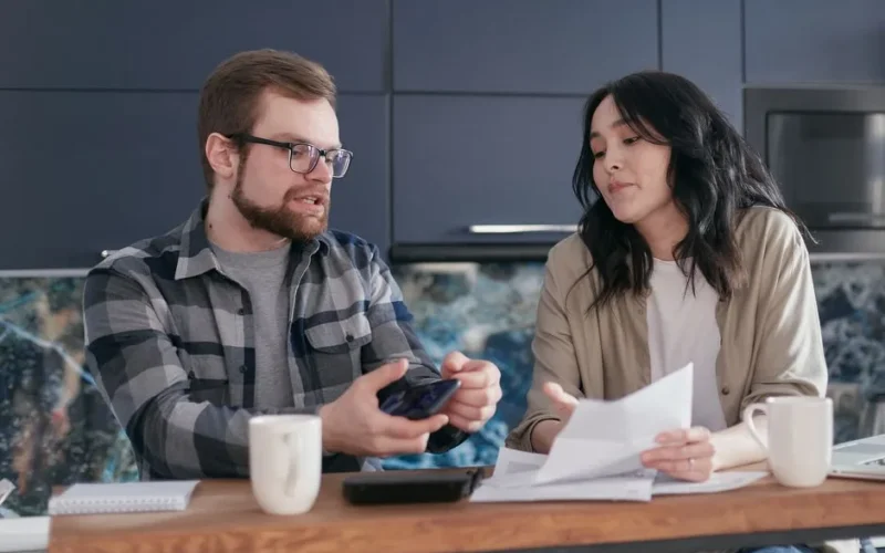 A couple sits at a kitchen table reviewing documents and discussing finances.