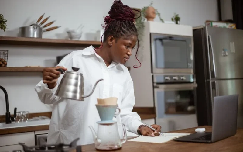A woman pours coffee into a filter while looking at a laptop in a chic kitchen.