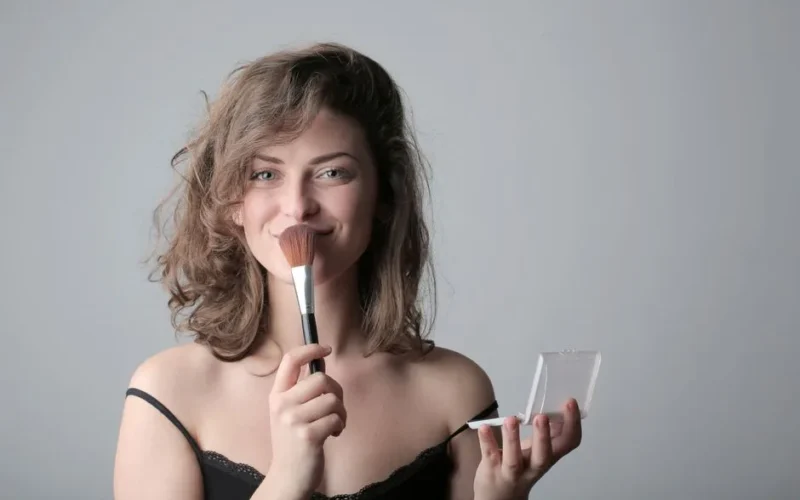 Smiling woman using a makeup brush in a studio setting, enhancing beauty with cosmetics.