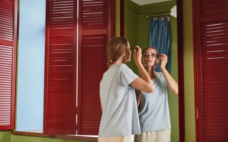 Woman applying mascara in front of a mirror with red shutters reflecting her image indoors.