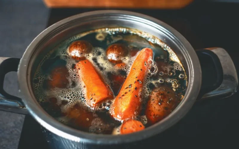 A close-up of carrots and potatoes boiling in a metal pot on a stove.