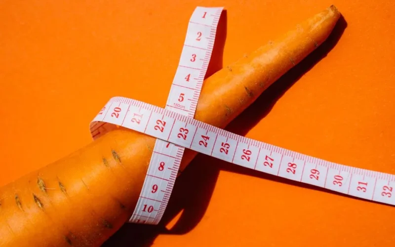 Close-up shot of a carrot wrapped with a measuring tape on a vibrant orange background, symbolizing healthy eating.