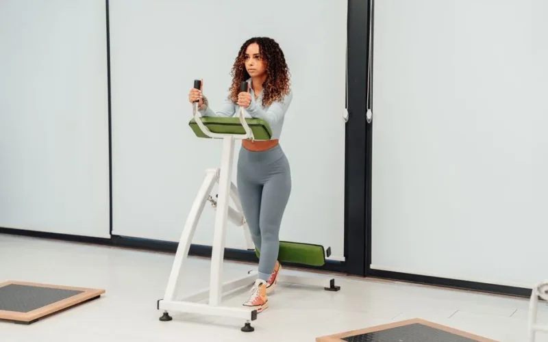 A woman with curly hair exercises on a machine in a bright gym.