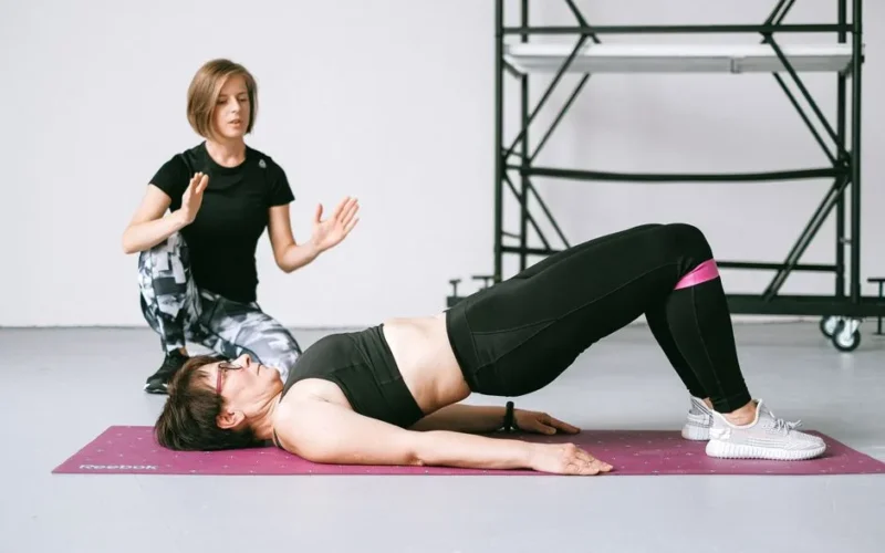 A personal trainer coaching a woman through a pelvic exercise on a yoga mat indoors.