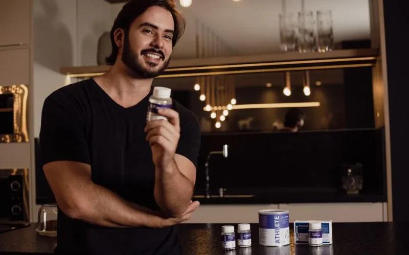 Smiling man holding a supplement bottle in an indoor kitchen setting with warm lighting.