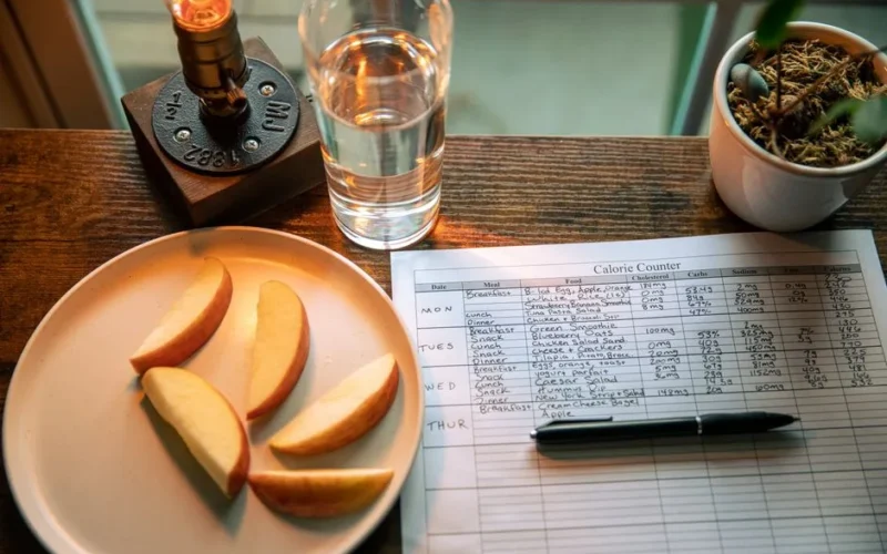A wooden table setup with apple slices, a calorie chart, and a glass of water for a healthy diet plan.