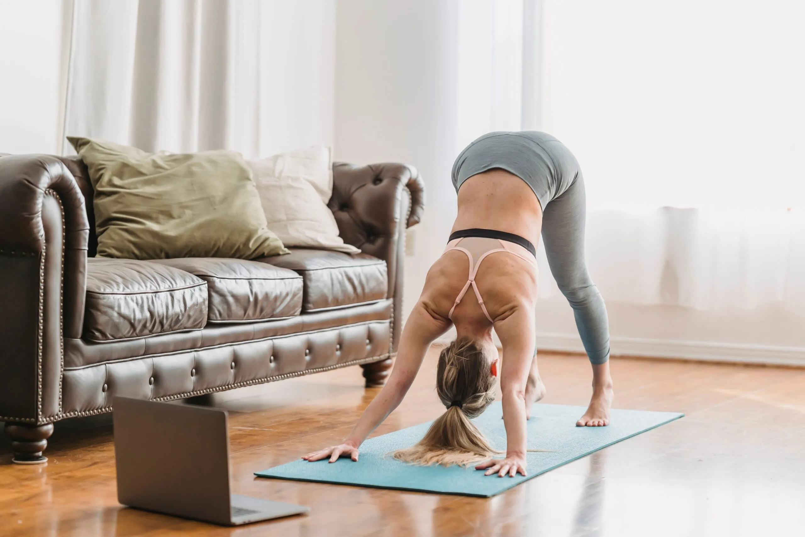 Full length of faceless lady wearing leggings and sportive bra stretching back on yoga mat near opened netbook and leather couch with pillows in light living room near curtains