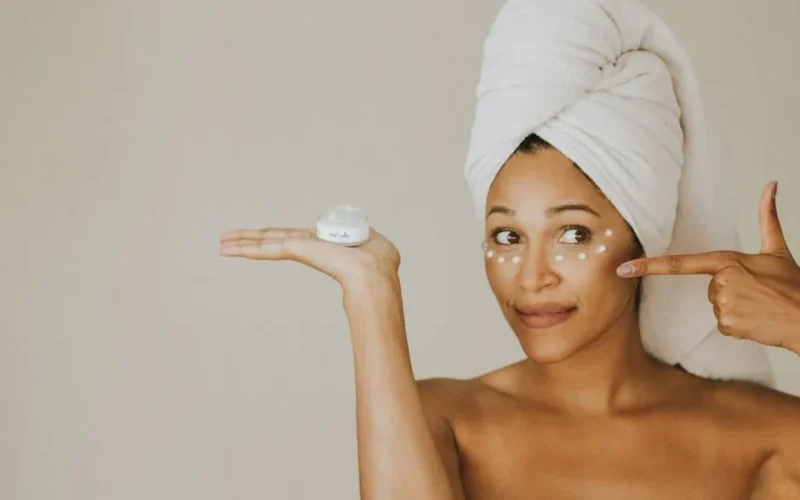 A woman applies under eye cream, holding a jar, with a towel on her head.