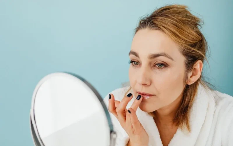 A woman in a bathrobe applies lip balm while looking in a mirror, highlighting skincare and self-care.