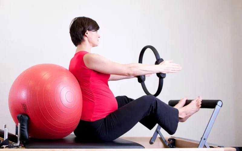 A pregnant woman in a red top practicing Pilates using a fitness ring and exercise ball in a gym setting.