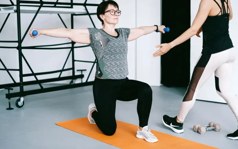 Two women working out with dumbbells on yoga mats in a modern gym setting, focusing on strength training.